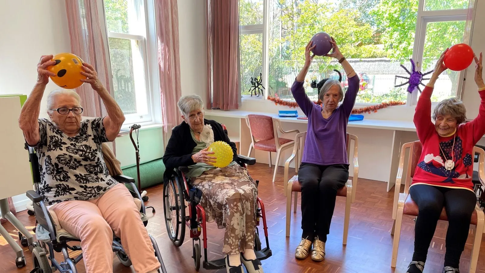 Older women exercising with small balls, seated in chairs and wheelchairs